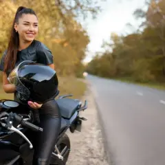 Uśmiechnięta kobieta w skórzanym stroju trzyma kask obok czarnego motocykla. Idealne motory dla kobiet na jesienną przejażdżkę.