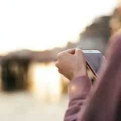 Osoba sprawdza na telefonie, jak włączyć roaming w Orange na kartę, podczas zachodu słońca nad wodą.