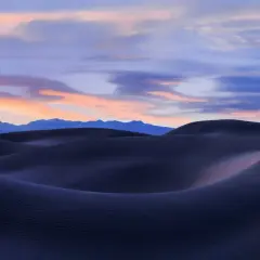 Landscape photography sunset over dark sand dunes, with soft pink and blue clouds painting the sky above distant mountains.