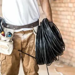 Elektryk z narzędziami w pasie trzyma zwój czarnego kabla. Czy będzie układany w peszlu, czy bezpośrednio w ziemi?