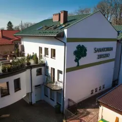 Sanatorium Zbysko w Busku-Zdroju, biały budynek z zielonym dachem i logo drzewa. Na tarasie wypoczywają kuracjusze.