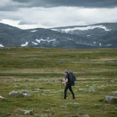 Vandrare med ryggsäck och stavar går över kalfjäll med snöfläckar på berg i bakgrunden. En helags karta skulle visa vägen.