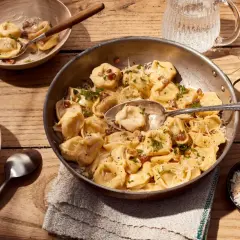 Herzhafte Tortellini Carbonara, garniert mit Petersilie und Parmesan, serviert in einer Pfanne und Schalen auf Holztisch.