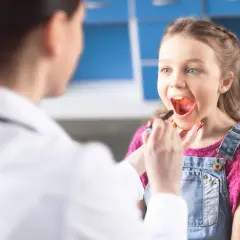 Lekarz bada gardło dziewczynki, która otwiera buzię. Słychać jej matowy głos, gdy mówi "aaa".