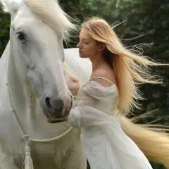 Una joven rubia con un vestido blanco se inclina hacia un majestuoso caballo blanco. Es como soñar con caballos, una escena etérea.