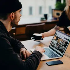 Mężczyzna w czapce pracuje na laptopie, analizując dane. Obok stoi filiżanka kawy. To praca ux researchera.