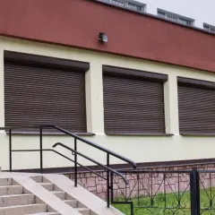Brązowe rolety nadstawne na jasnym tynku budynku, z widocznymi schodami i barierkami.