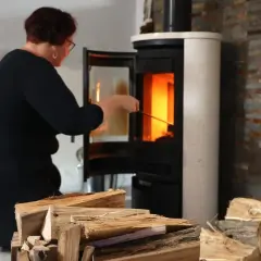 Frau kniet vor einem brennenden Kamin, um ihn zu reinigen. Holzscheite liegen bereit.
