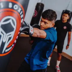 A young boxer practices his boxing combos, throwing a punch at a heavy bag in a gym. Another person watches in the background.
