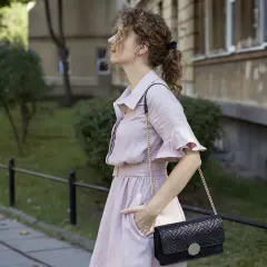 Kobieta w różowej sukience i czarnej torebce na łańcuszku na tle budynku i zieleni.