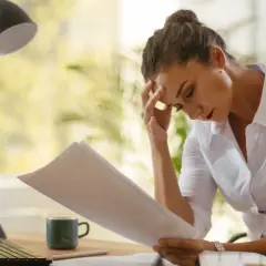 Frau mit Kopfschmerzen blickt auf Dokumente, die sie vielleicht für eine verbindliche Auskunft beim Finanzamt benötigt.