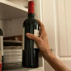 Dłoń sięga po butelkę bordeaux wina z półki, obok stosu talerzy i miseczek.