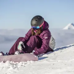 Kobieta w kasku i goglach siedzi na śniegu, zapinając deskę snowboardową.