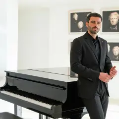 Uomo elegante in completo nero, pronto per un'esibizione teatrale, accanto a un pianoforte a coda.