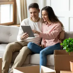 Couple sur un canapé, regardant une tablette. Des cartons de déménagement sont autour d'eux, peut-être pour une lettre pour quitter son logement sans préavis.