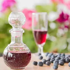 Elegancka karafka z nalewką z borówki, obok kieliszek i garść świeżych owoców. W tle kwitnące kwiaty.