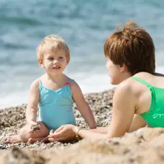 Mama i dziecko cieszą się wakacjami nad morzem z dziećmi. Dziewczynka w niebieskim kostiumie bawi się kamyczkiem, a mama w zielonym leży obok.