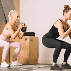 Dwie kobiety wykonujące przysiady z kettlebellami w siłowni.