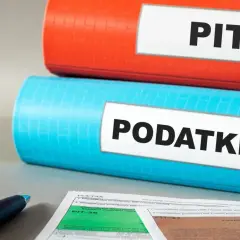 Dwa segregatory, jeden czerwony z napisem "PIT", drugi niebieski z napisem "PODATKI", leżą na biurku obok formularzy podatkowych i długopisu.