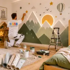 Maler gestaltet Kinderzimmer Wandgestaltung mit Bergen, Sonne und Sternen. Ein Heißluftballon schwebt über der Szene.