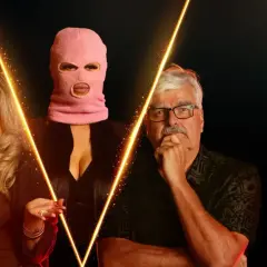 Tre personer, en med rosa balaklava, står framför en gul ljusformation. En kvinna till vänster, en man med glasögon till höger. Kanske från spelet SVT.
