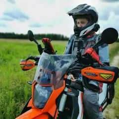 Motocyklista w kasku siedzi na pomarańczowym motocyklu w terenie, na tle zielonego pola i nieba.