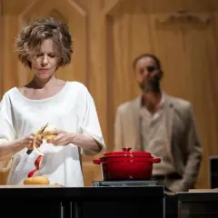 La locandiera recensioni: donna sbuccia una mela, pentola rossa sul fuoco, uomo sullo sfondo.