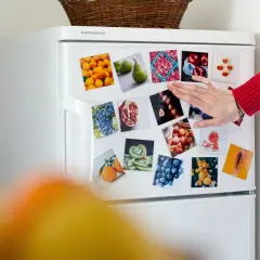 Biała lodówka z magnesami przedstawiającymi owoce. Dłoń w czerwonym swetrze dotyka jednego z magnesów.