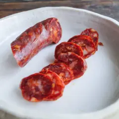 Ein Stück und Scheiben einer würzigen Salsiccia Wurst liegen in einer weißen Schale auf einem Holztisch.