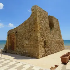 Antica torre di pietra sulla costa, con il mare azzurro sullo sfondo. Vicino, una fila di vasi di terracotta, come tre fontane in attesa.