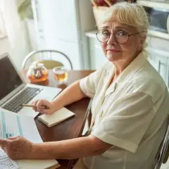 Starsza kobieta z okularami analizuje dokumenty, zastanawiając się, ile wynosi emerytura matczyna. Obok laptop, herbata i notatnik.