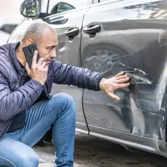 Mężczyzna sprawdza uszkodzenia na samochodzie po stłuczce samochodowej nie z jego winy. Dzwoni po pomoc, wskazując na głębokie rysy.