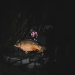 Wędkarz z dumą prezentuje ogromnego karpia, którego udało mu się złowić nocą. To niezapomniane chwile, kiedy biorą ryby.