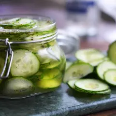 A jar of sliced pickled cucumbers in brine, with more slices scattered on a marble surface.