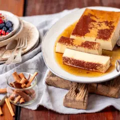 Pyszny creme caramel na talerzu, polany syropem. Obok miseczka z owocami i laska cynamonu.