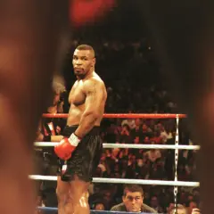 Mike Tyson full body, standing in a boxing ring, wearing red gloves and black shorts, with a crowd watching.