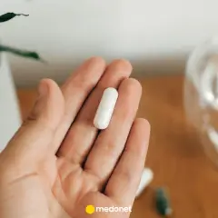 Dłoń trzyma białą kapsułkę. Obok szklanka wody. Uważaj na nadmiar magnezu objawy, by nie przedawkować.