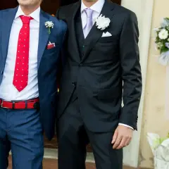 Dwóch mężczyzn w garniturach, jeden z nich to świadek na ślubie, ubrany w niebieski garnitur w kratę i czerwoną krawat w kropki.