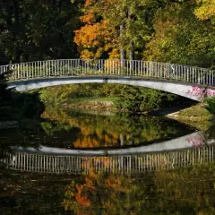 Mostek nad spokojną rzeką, odbijający jesienne barwy drzew. W oddali widać ludzi spacerujących po moście.