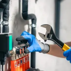 Instalator w niebieskich rękawiczkach reguluje mieszacz wody ciepłej i zimnej za pomocą klucza.