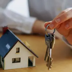 Mano sostiene llaves frente a maqueta de casa. Una posible solución si un heredero no quiere vender.