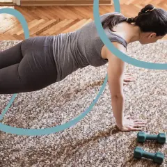 Kobieta w pozycji deski ćwiczy pilates w domu, patrząc na laptopa. Obok leżą hantle.