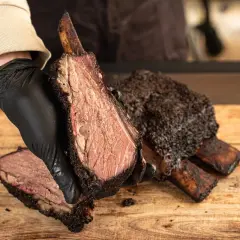 Une main gantée présente une côte de bœuf fumée, tendre et juteuse, sur une planche en bois.