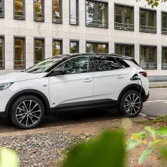 Biała hybryda plug-in ładuje się przy stacji ładowania. Nowoczesny samochód stoi na tle budynku z wieloma oknami.