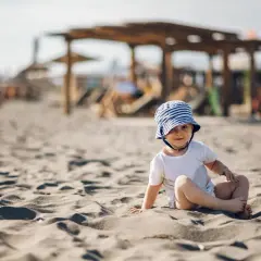 Małe dziecko w pasiastym kapeluszu siedzi na piaszczystej plaży, w tle widać parasole i leżaki.