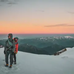 Deux alpinistes entament leur ascension du Mont Rose au lever du soleil, le ciel aux teintes roses et orangées embrassant les sommets enneigés.