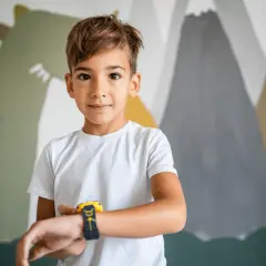 Chłopiec w białej koszulce z zegarkiem na ręku, na tle kolorowej ściany z motywem gór i dinozaura. Może to być inspiracja dla imiona dla chłopców.