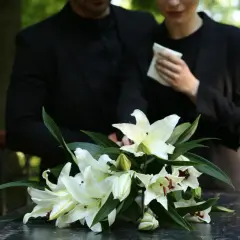 Białe lilie na ciemnym blacie, w tle para w żałobie.