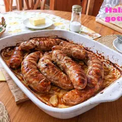 Pyszna, pieczona kiełbasa biała w naczyniu żaroodpornym, z cebulką i czosnkiem. Obok koszyk z jajkami i kromka chleba.
