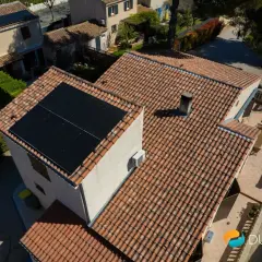 Toits de maisons avec panneaux solaires noirs. Une unité extérieure de pompe à chaleur est visible sur le mur.